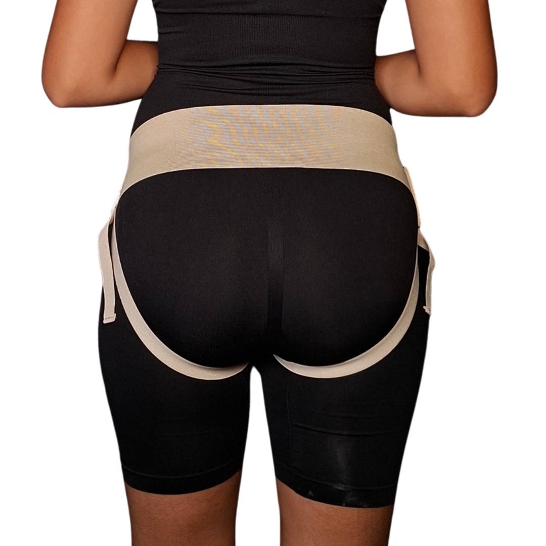 Vista frontal y posterior de un braguero inguinal unisex en tono beige o crema colocado sobre prenda ajustada negra. Muestra una banda pélvica elástica ancha con sistema de ajuste mediante broche plástico y correas perineales ajustables que pasan por debajo de la zona glútea para brindar compresión localizada en la región inguinal.